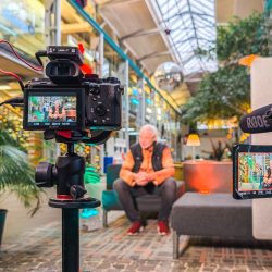 Ein Mann sitzt auf einem Sofa in einem modernen Innenraum, während er von zwei Kameras auf Stativen gefilmt wird; die Kamerabildschirme zeigen ihn im Bild.