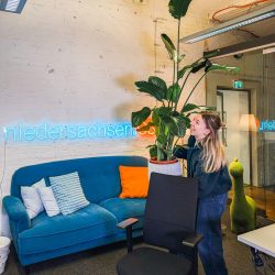 Eine Frau steht in einem Büro neben einer großen Topfpflanze, mit einer blauen Couch, einem Neonschild "niedersachsen.de" und einem Bürostuhl im Hintergrund.