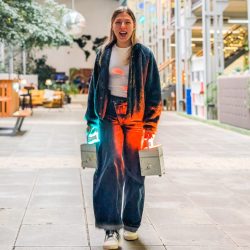 Eine junge Frau geht in einem Haus auf einem gefliesten Boden und hält zwei kleine Schachteln mit leuchtenden Lichtern, eine rote und eine blaue, in jeder Hand. Sie lächelt und trägt legere Kleidung.