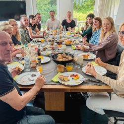 Eine Gruppe von Menschen sitzt in einem gut beleuchteten Raum um einen Esstisch mit Tellern voller Essen, Getränken und Frühstücksartikeln herum, lächelt und blickt in die Kamera.