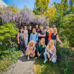 Eine Gruppe von dreizehn Menschen posiert und lächelt in einem üppigen, grünen Garten mit blühenden Glyzinien und hellem Sonnenlicht.