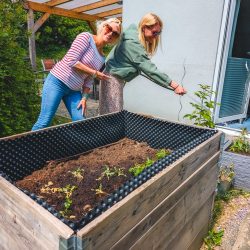 Zwei Frauen pflegen draußen in der Nähe eines Hauses ein erhöhtes Holzbeet mit jungen Pflanzen.