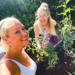Zwei Frauen sind bei der Gartenarbeit im Freien, lächeln in die Kamera, die Sonne scheint auf sie und grüne Pflanzen umgeben sie.