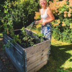Eine Frau gießt an einem sonnigen Tag Pflanzen in einem Hochbeet aus Holz und lächelt in die Kamera.