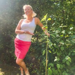 Eine Frau in einem weißen Tanktop und einem rosa Rock steht im Freien, lächelt und deutet auf eine hohe Grünpflanze in einem Garten.