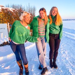 Drei Frauen in grünen Pullovern stehen zusammen und lächeln auf einem verschneiten Weg unter einem klaren blauen Himmel.