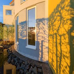 Gebogene Außenwand eines modernen Hauses mit großen Fenstern, Sonnenlicht wirft Baumschatten auf gelbe und graue Fassaden, Felsen und ein Teich im Vordergrund.