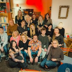 Eine Gruppe von achtzehn Personen sitzt und steht in einem gemütlichen, gut beleuchteten Raum mit Bücherregalen und Dekoration zusammen.