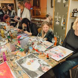 Eine Gruppe von Menschen sitzt an einem Tisch, der mit Malutensilien und Gemälden bedeckt ist, und ist in einem hell erleuchteten Atelier damit beschäftigt, Kunstwerke zu schaffen und sich zu unterhalten.