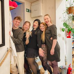 Fünf Personen stehen in einem Haus, lächeln und posieren zusammen in der Nähe einer Treppe und einer Wand, an der im Hintergrund ein Feuerlöscher und grüne Fluchtwegschilder zu sehen sind.