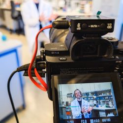 Eine Kamera filmt eine Person in einem Laborkittel, die in einem Labor steht; das Kameradisplay zeigt ein klares Bild der Person und der Laborausrüstung.