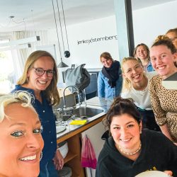 Eine Gruppe von acht Personen, meist Frauen, lächelt gemeinsam in einer modernen Küche; eine Person im Vordergrund hält einen Teller mit einem runden Stück Brot.