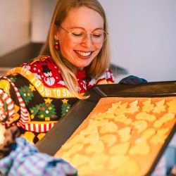 Eine Frau trägt einen Weihnachtspullover und hält ein Tablett mit Keksen.