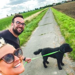 Zwei Menschen gehen mit einem schwarzen Hund an der Leine einen ländlichen Weg entlang, mit Feldern und einem bewölkten Himmel im Hintergrund.